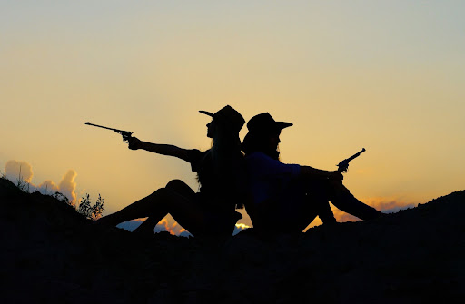 Two people holding guns sitting back to back against a sunset. 